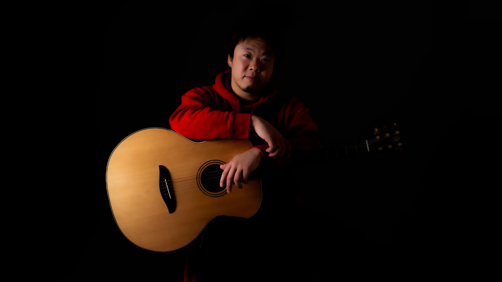 Masashi Hasegawa seated with an acoustic guitar against a dark background.